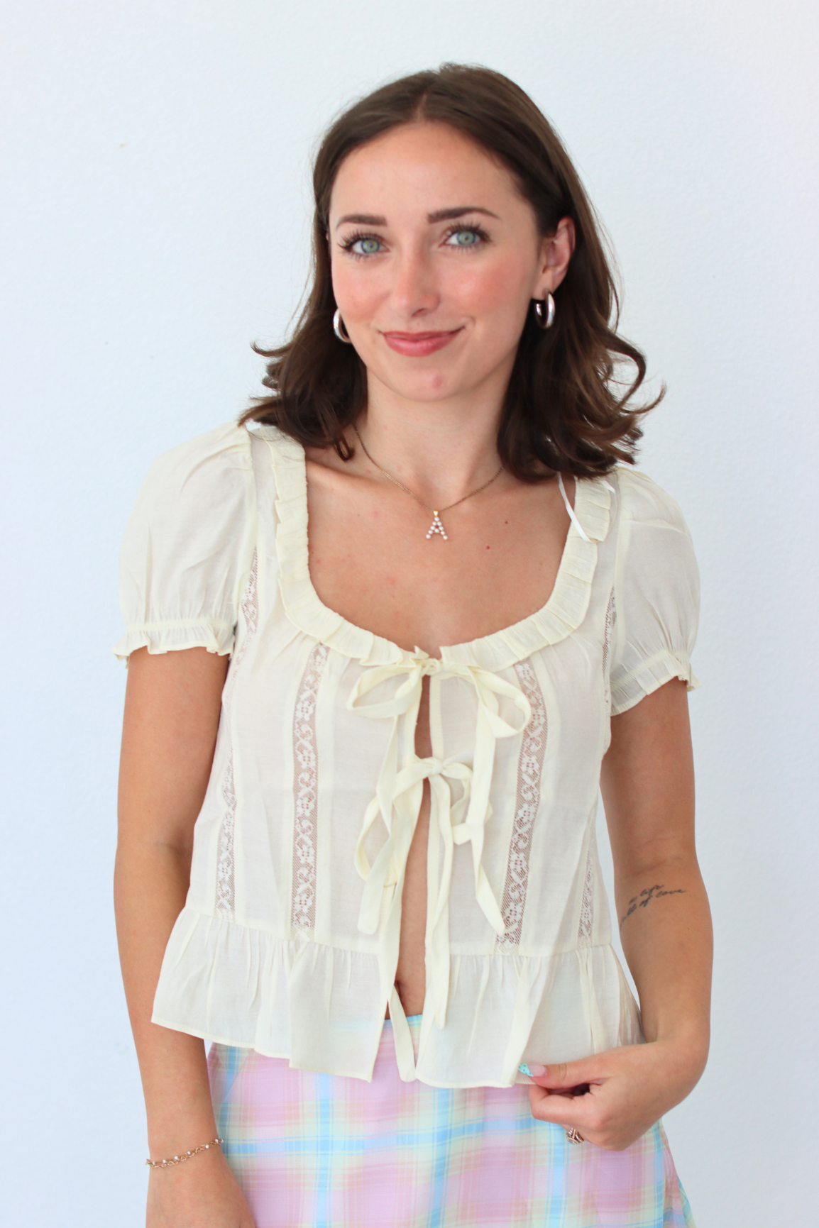 A woman smiling and posing in a cream-colored short-sleeve blouse with lace trim and a tie-front detail, paired with a pastel plaid skirt, against a plain light background.