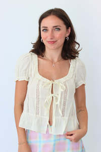 A woman smiling and posing in a cream-colored short-sleeve blouse with lace trim and a tie-front detail, paired with a pastel plaid skirt, against a plain light background.