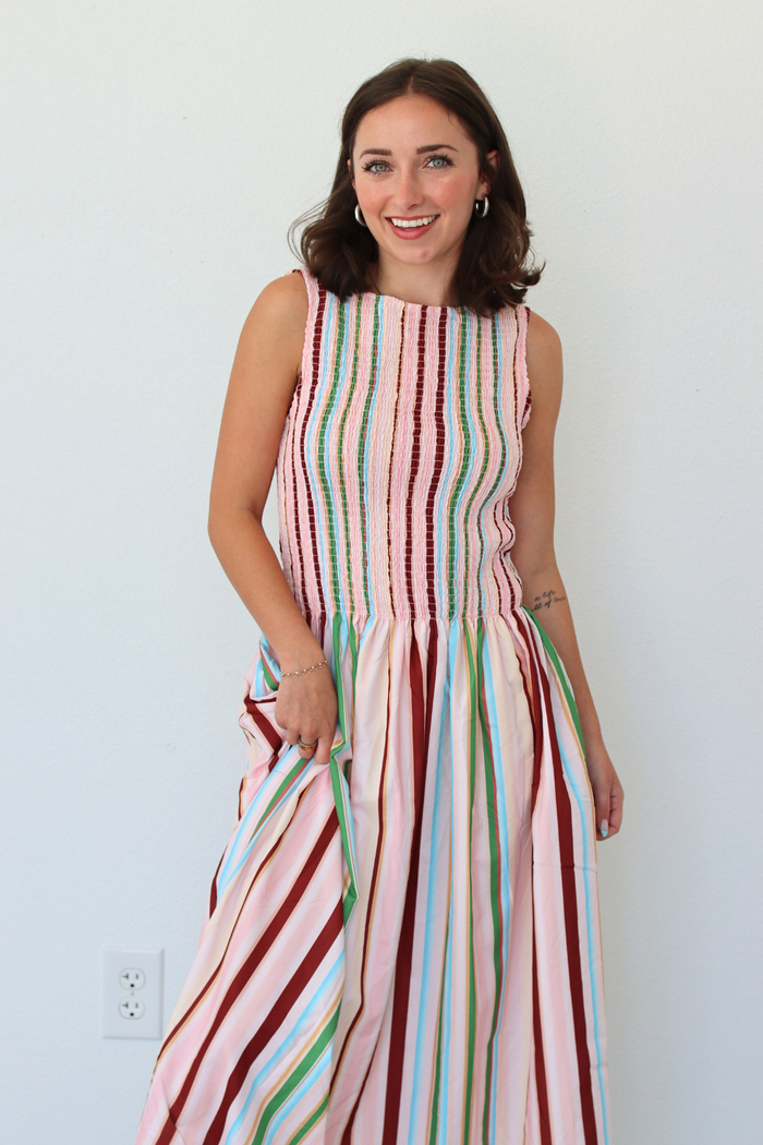 A woman wearing a sleeveless, smocked maxi dress with vertical multicolor stripes in pink, red, green, blue, and white, standing against a plain white wall.