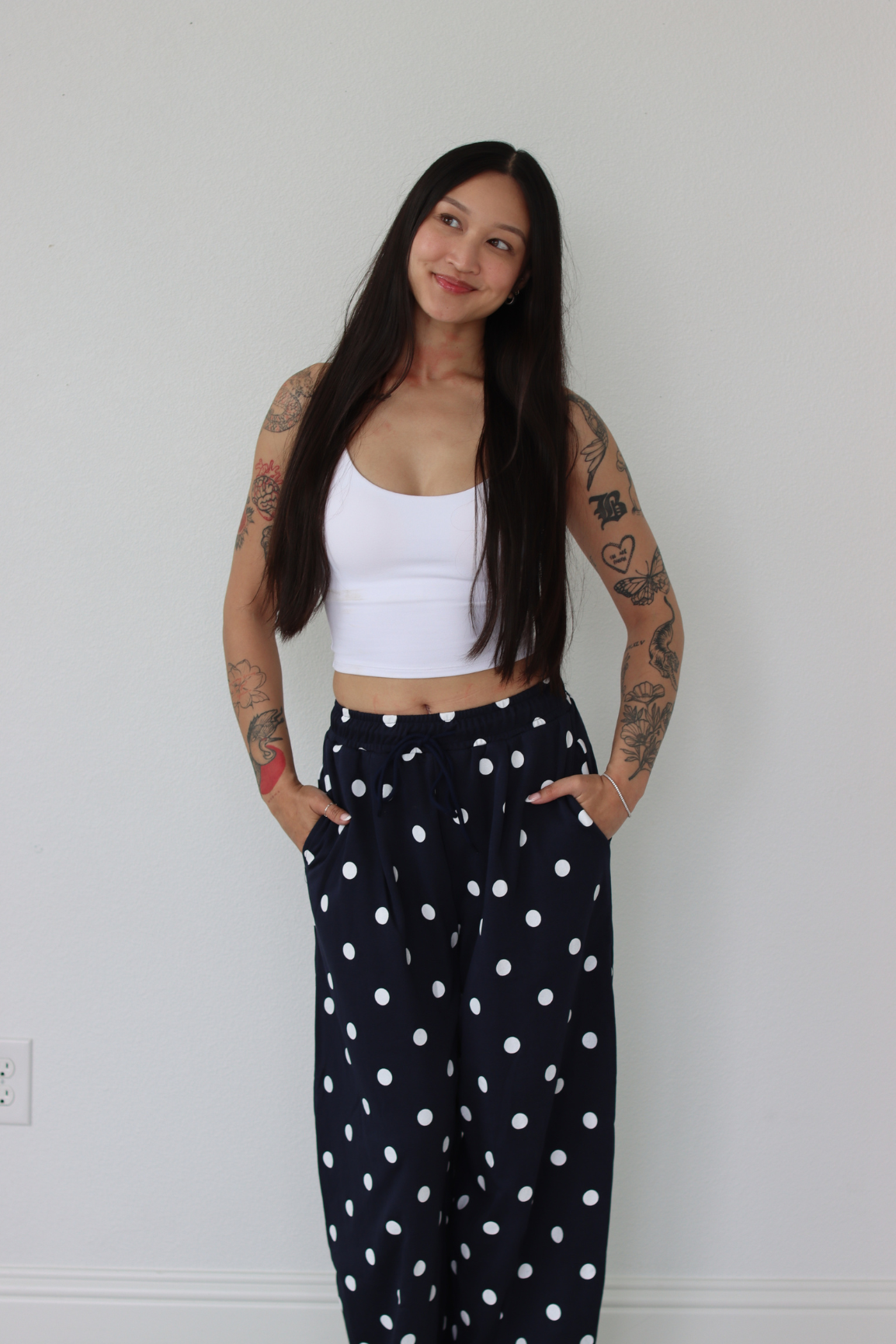 Woman wearing a white top and navy polka dot pants against a white wall.