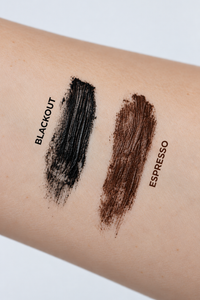 Close-up of a forearm showing two makeup swatches: a black textured swipe labeled BLACKOUT and a medium brown textured swipe labeled ESPRESSO.