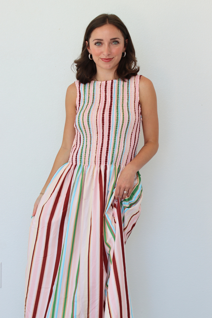 A woman modeling a sleeveless maxi dress with vertical multicolor stripes in pink, red, green, and blue on a light gray background.