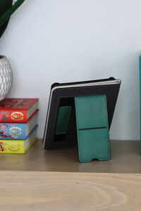 A kindle case with a teal green front cover standing open on a wooden shelf, next to a stack of colorful books against a light wall.