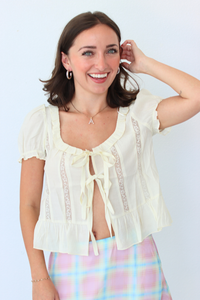 A woman smiling and posing in a cream-colored short-sleeve blouse with lace trim and a tie-front detail, paired with a pastel plaid skirt, against a plain light background.