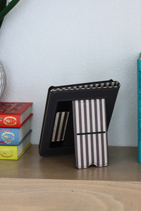 A brown leather kindle case with a striped card kickstand, standing open on a wooden shelf next to books against a light blue wall.