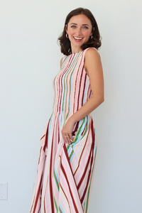 A woman modeling a sleeveless maxi dress with vertical multicolor stripes in pink, red, green, blue, and white against a plain white wall.