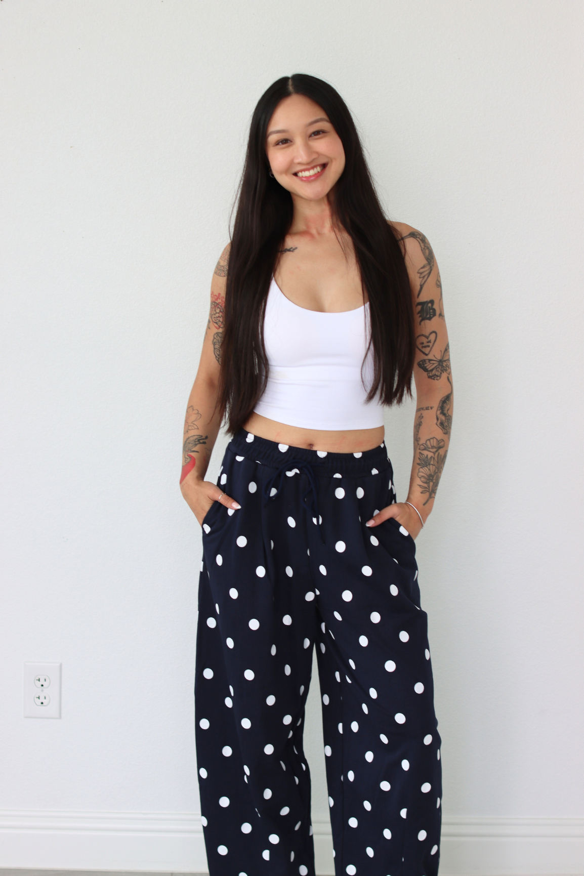 Woman wearing a white top and navy polka dot pants against a white wall.
