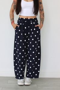 Woman wearing a white top and navy polka dot pants against a white wall.