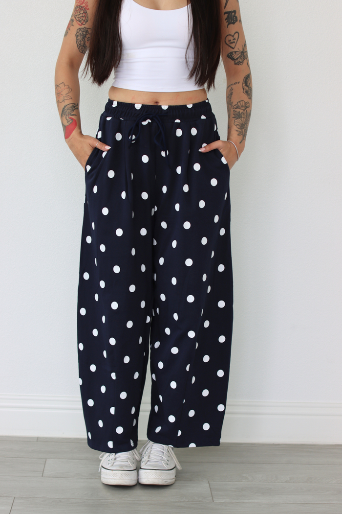 Woman wearing a white top and navy polka dot pants against a white wall.