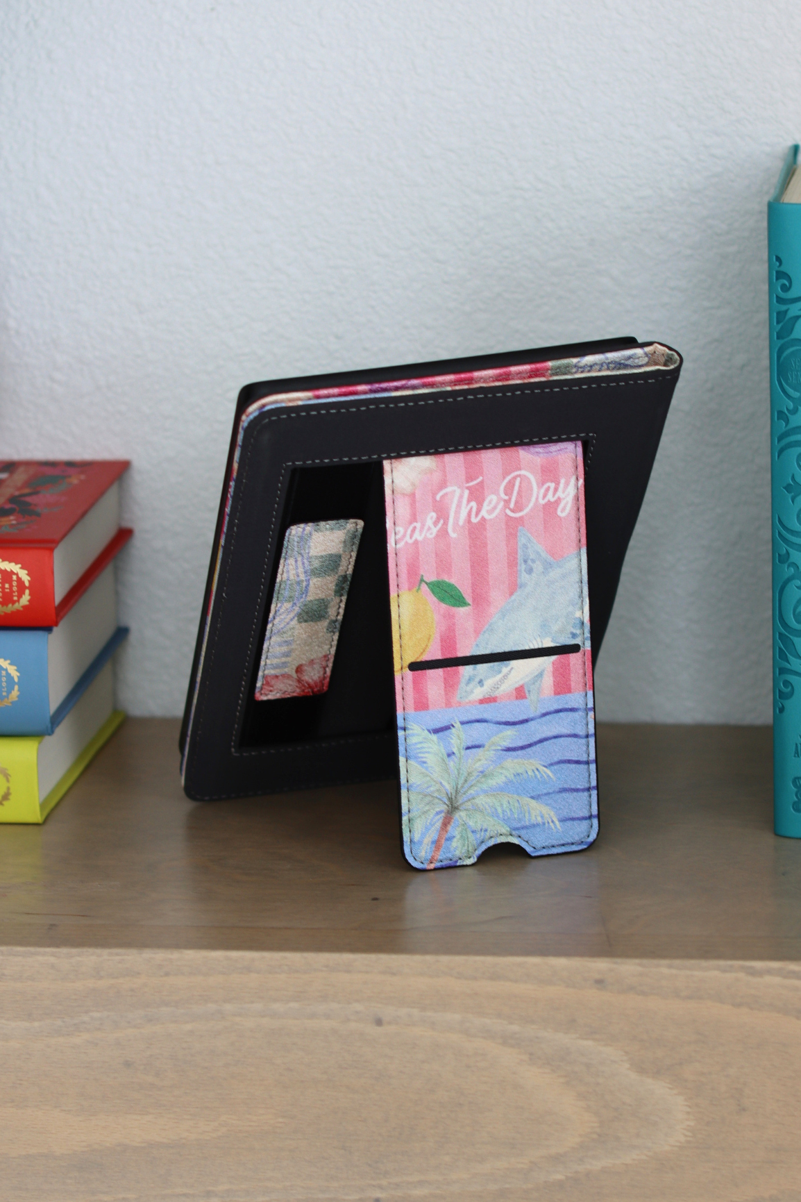 A kindle case standing on a wooden shelf, holding a colorful patterned phone case. The case has a pink and blue tropical beach design with the words 'Be The Day'.