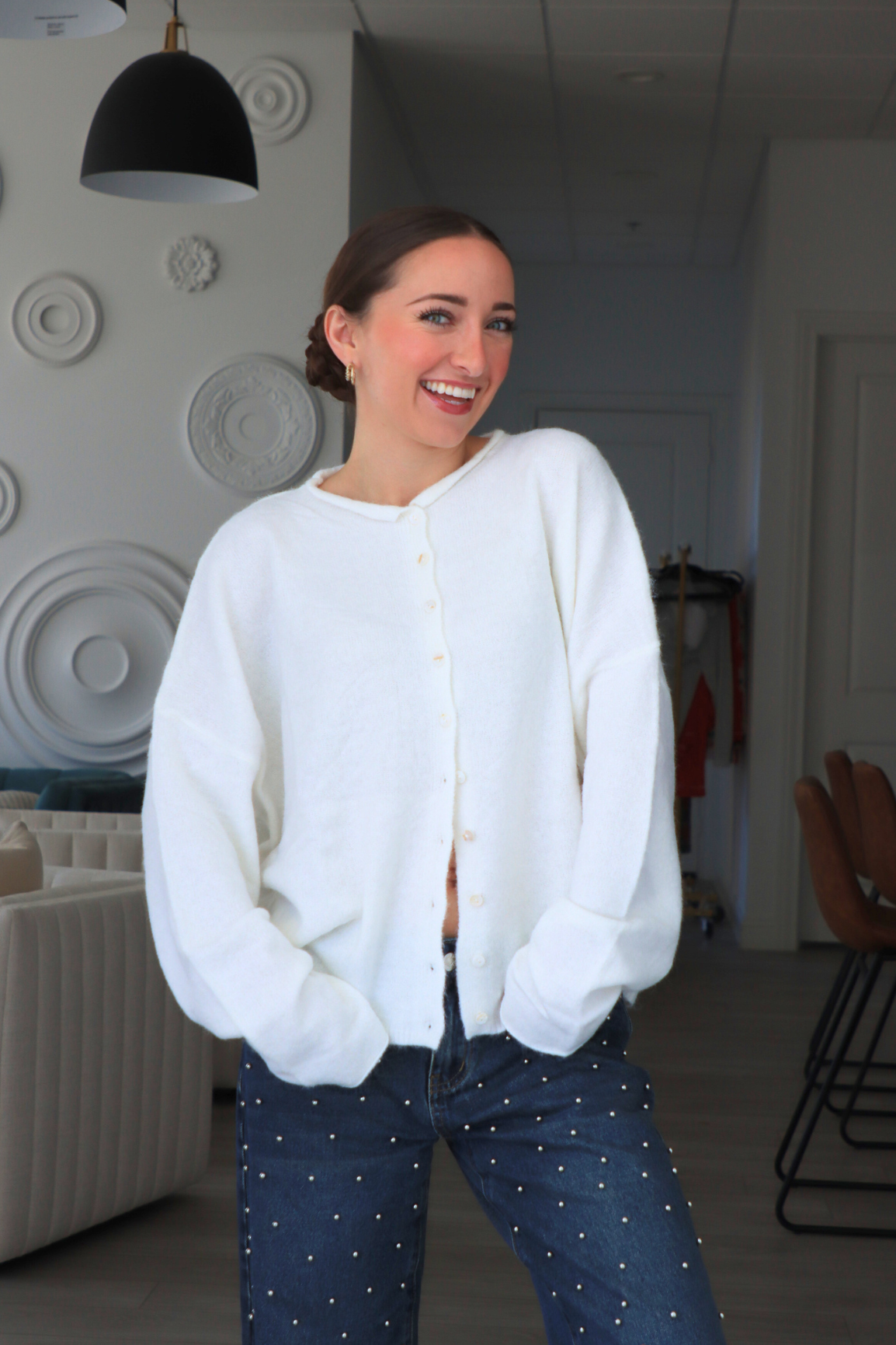 Woman wearing a white cardigan and blue jeans indoors.