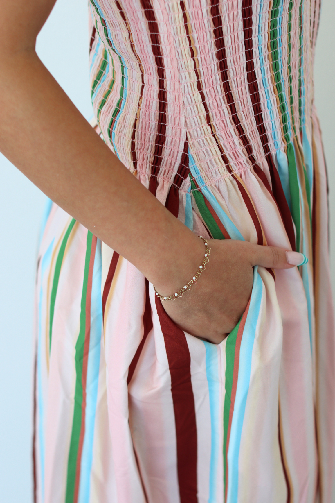 A close-up of a person wearing a pastel striped dress with vertical bands of pink, cream, green, blue, and burgundy. The person's hand is tucked into the side pocket of the dress, and a delicate beaded bracelet is visible on the wrist.