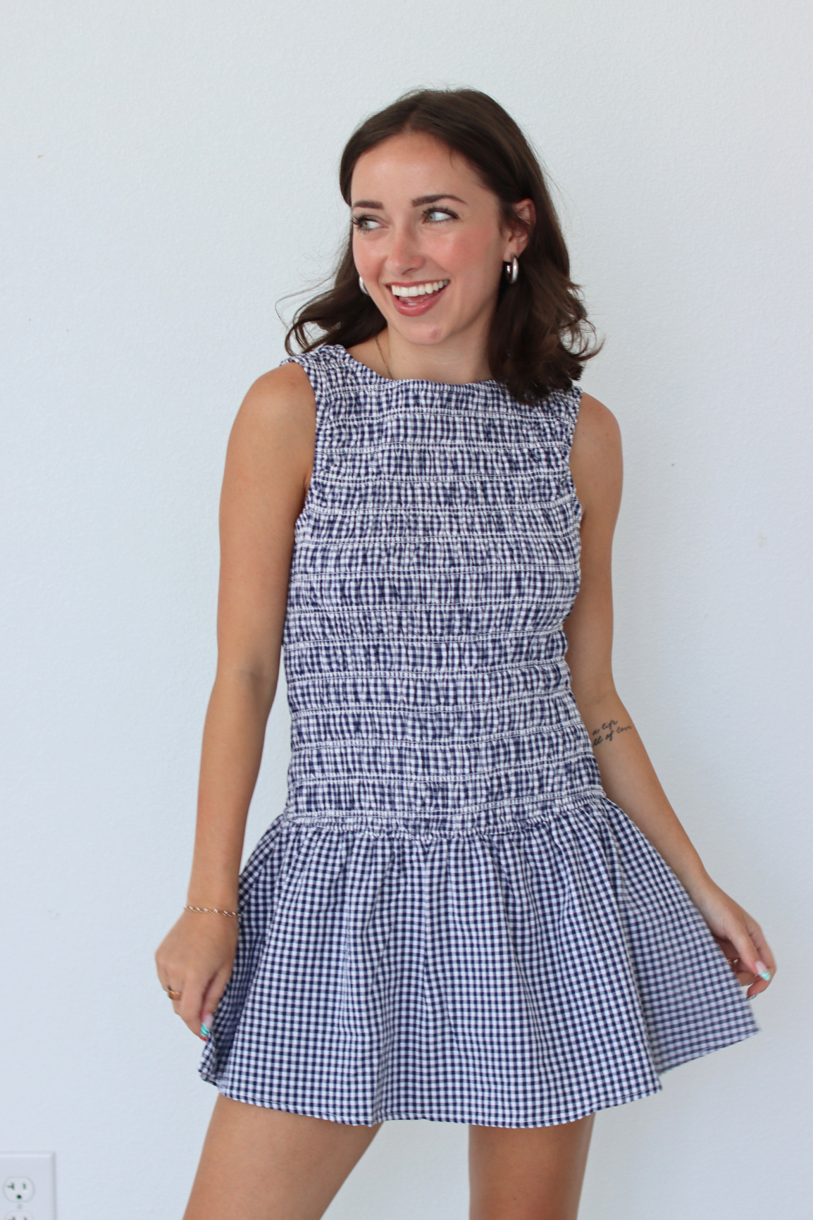 A woman modeling a sleeveless smocked mini dress with a blue and white gingham pattern, standing against a plain white wall.