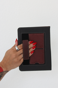 A hand with red nail polish and silver rings holds a kindle case with a burgundy kickstand and hand strap against a plain white background.