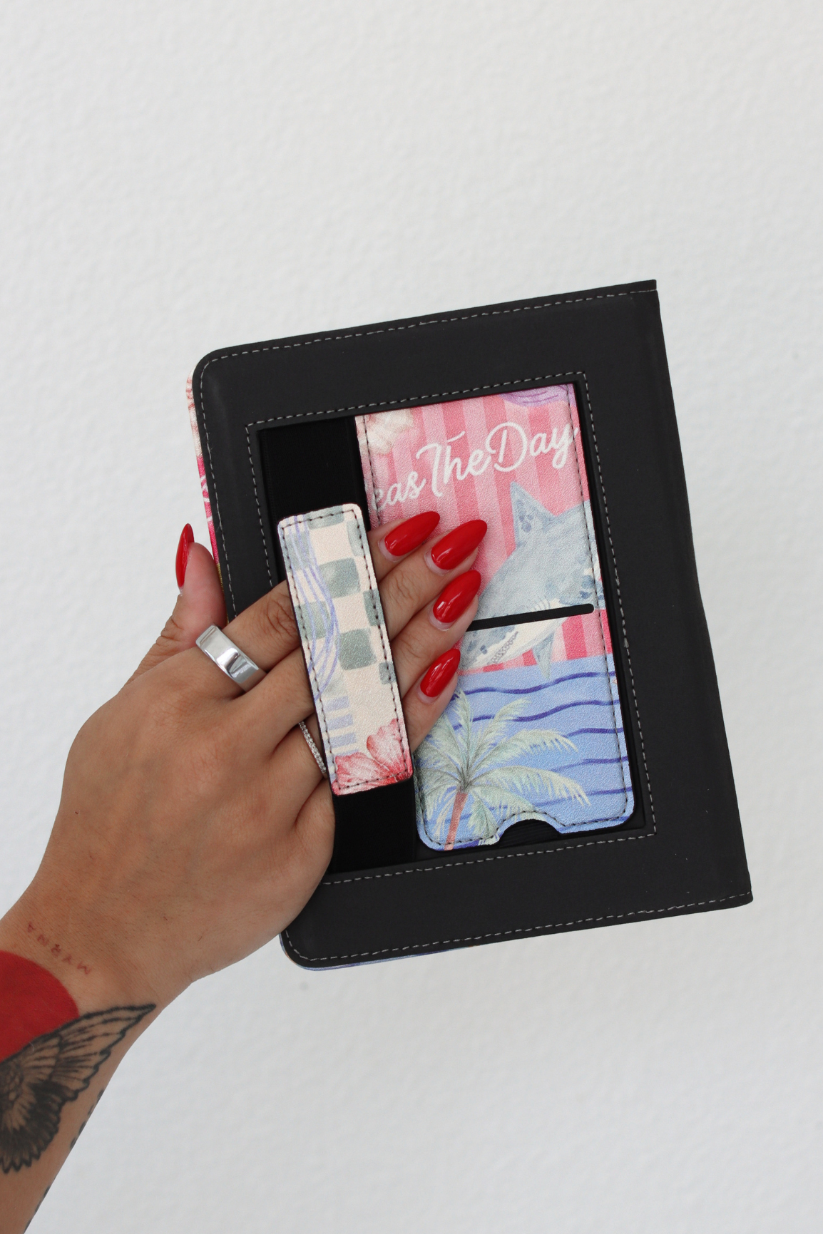 A hand with red nail polish holds a kindle case with colorful printed kickstand and hand strap, against a plain white wall