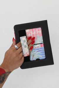 A hand with red nail polish holds a kindle case with colorful printed kickstand and hand strap, against a plain white wall
