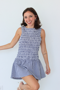 A woman modeling a sleeveless smocked mini dress with a blue and white gingham pattern, posing against a plain white wall.