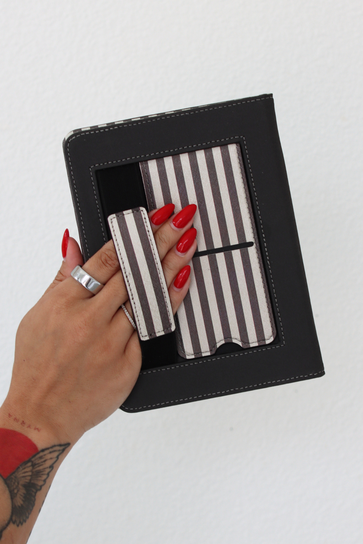 A hand with red nail polish holds a brown faux-leather kindle case containing striped hand strap and a matching striped kickstand, against a plain white background.