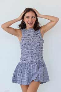 A woman modeling a sleeveless smocked mini dress with a blue and white gingham pattern, set against a plain white wall.