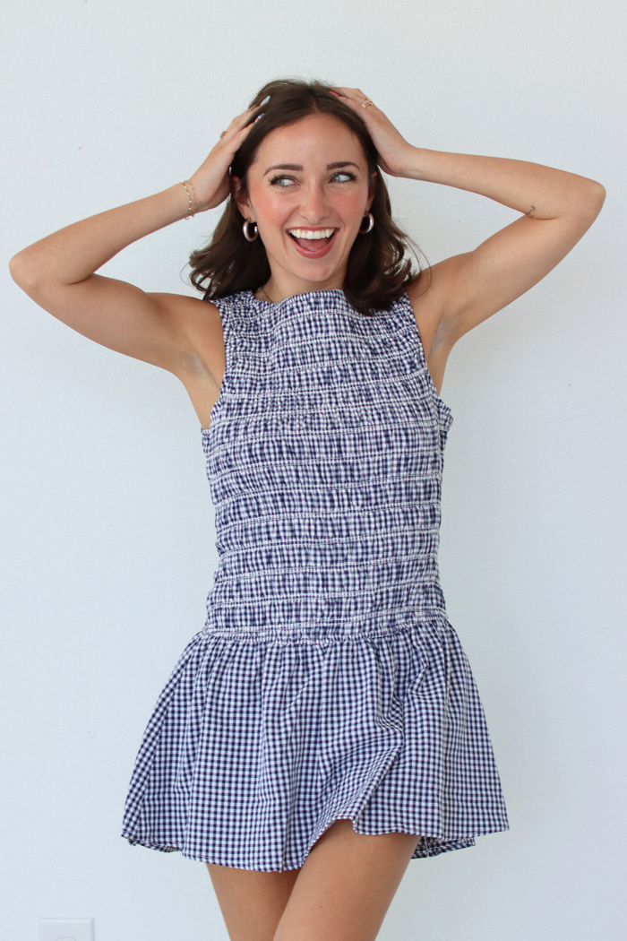 A woman modeling a sleeveless smocked mini dress with a blue and white gingham pattern, set against a plain white wall.
