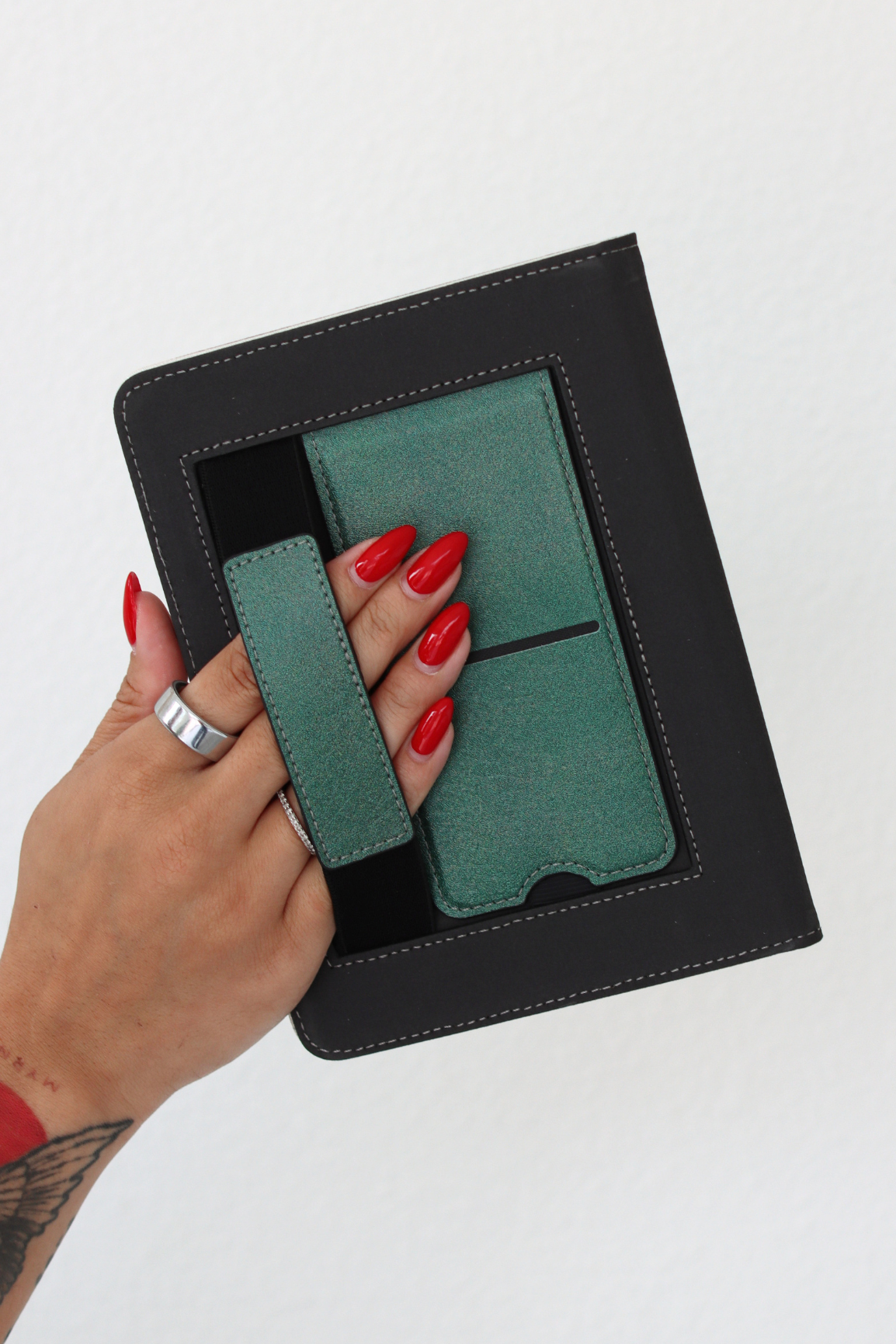 A hand with red manicured nails holds a kindle case with a green kickstand and hand strap against a plain white background.