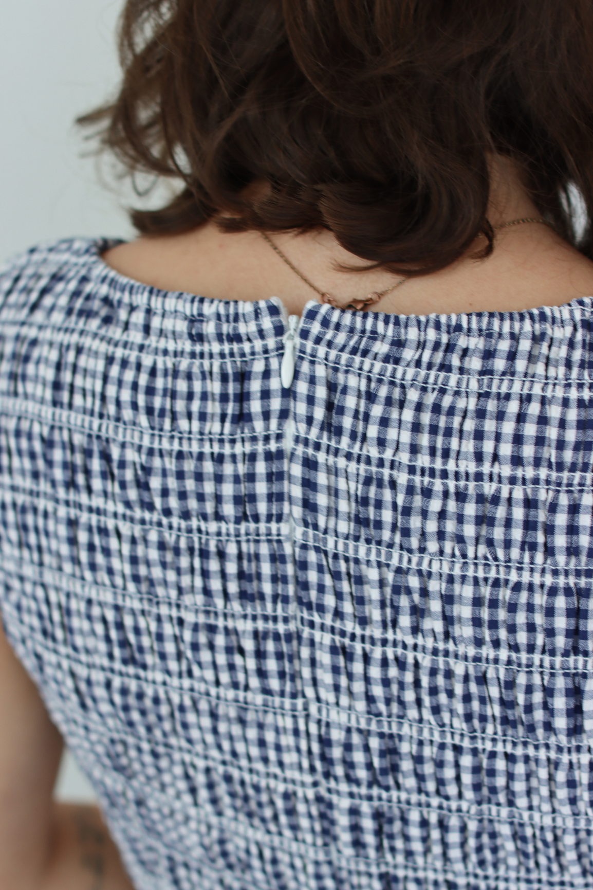 Close-up back view of a person wearing a smocked off-the-shoulder top with a navy and white gingham pattern.
