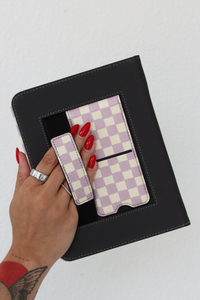 A hand with red nail polish holds a nook case against a white background, showing a light purple and cream checkered strap and kickstand