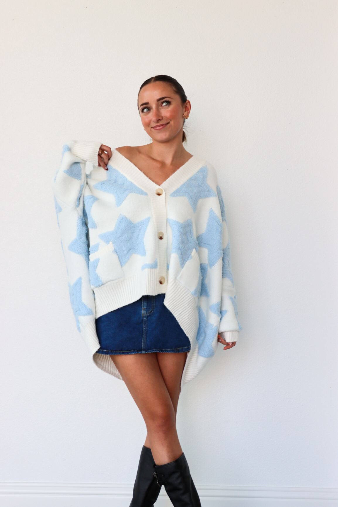 Woman wearing a blue and white patterned cardigan over a white shirt with a denim skirt and black knee-high boots.