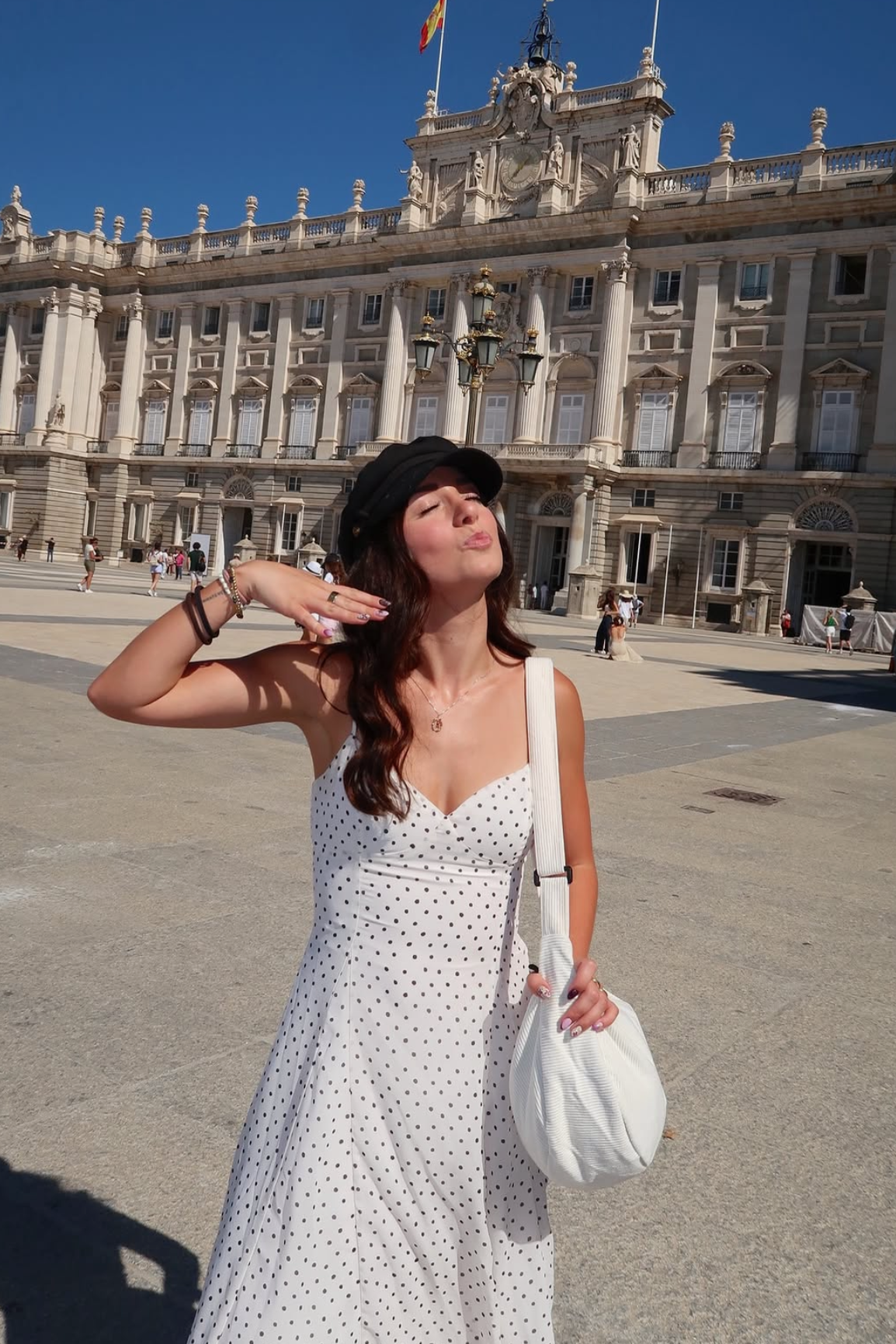 A woman posing outdoors in front of a grand classical building, wearing a white sleeveless dress with black polka dots, a black hat, and carrying a white shoulder bag.