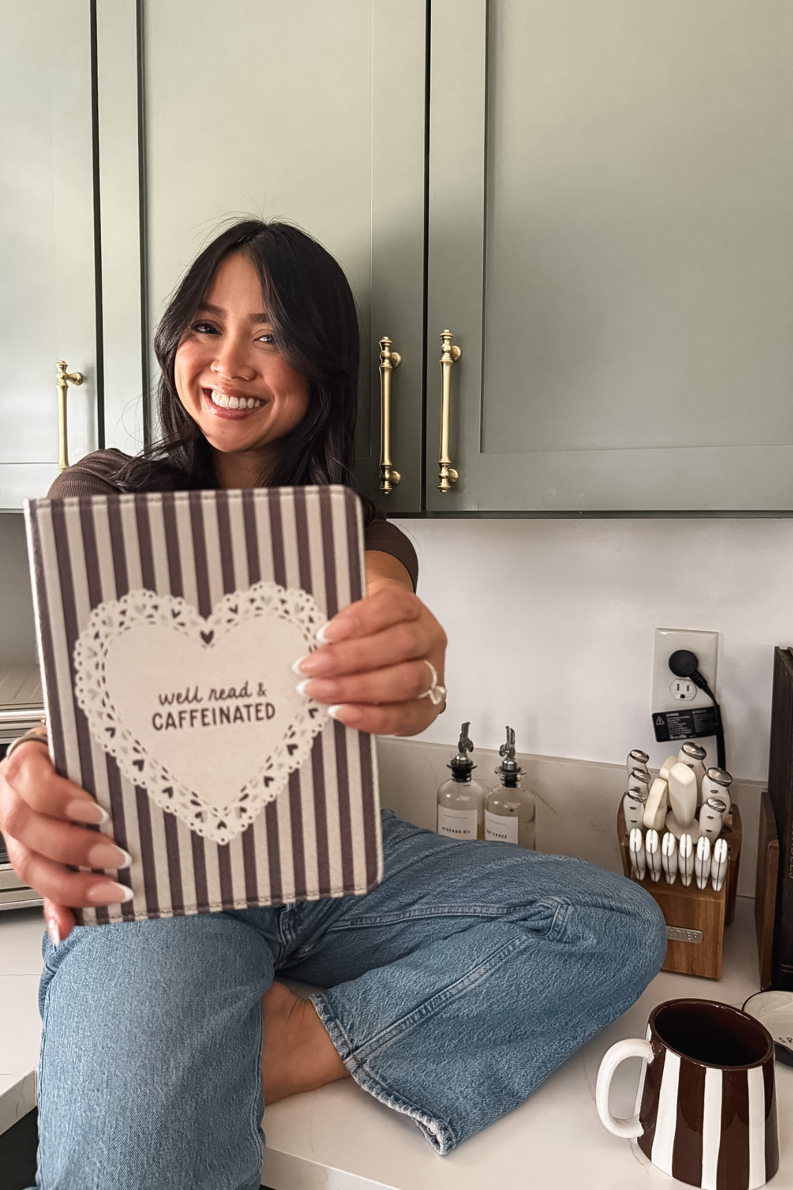A smiling woman sitting on a kitchen counter holds up a striped kindle case with a heart-shaped lace design and the text 'well read a CAFFEINATED'. The background shows sage green cabinets, gold handles, and a wooden knife block.