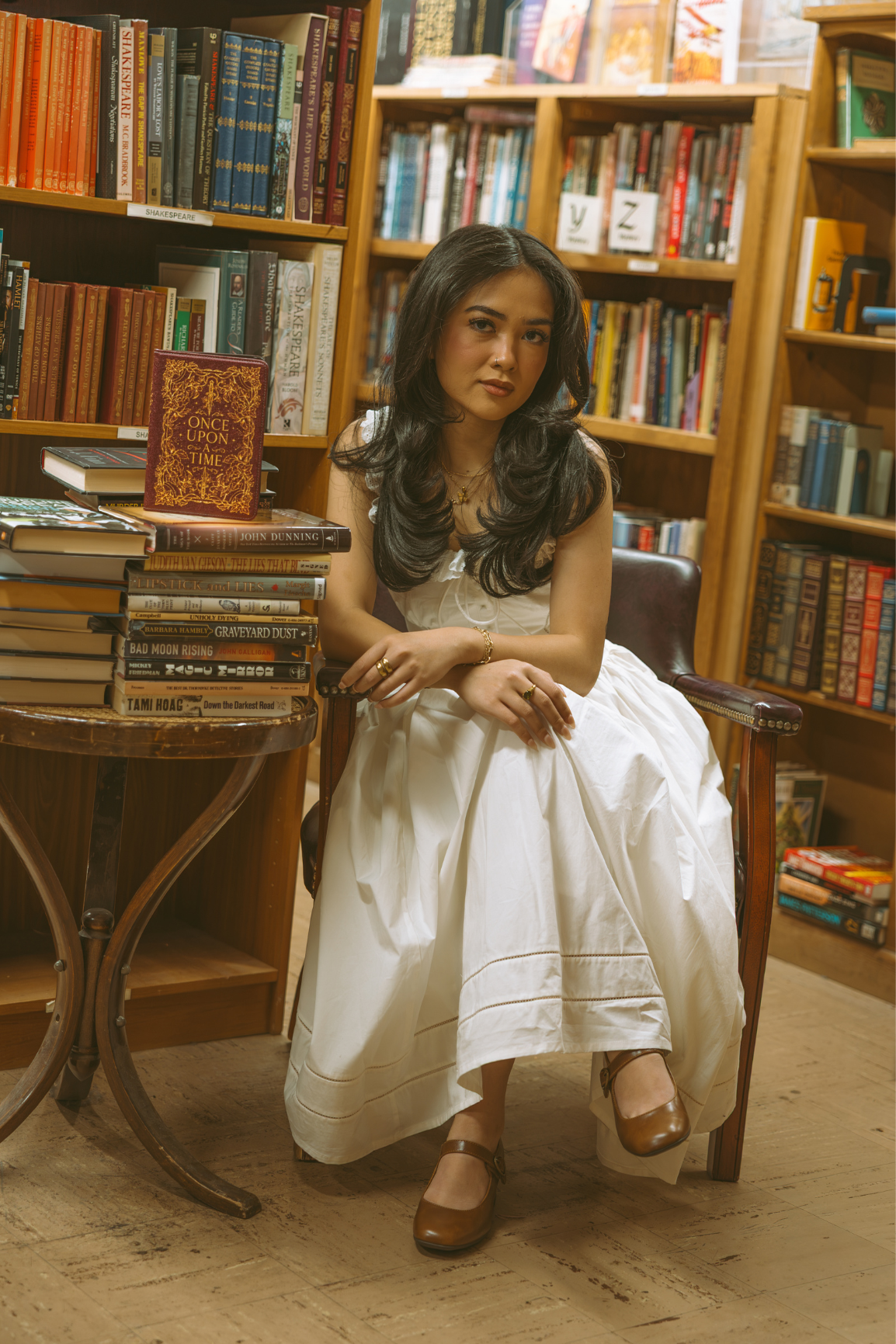 A woman with long dark hair sits in a wooden chair inside a book-filled room, wearing a white sleeveless dress and brown Mary Jane shoes. Wooden bookshelves and a small table with stacked books are visible around her.