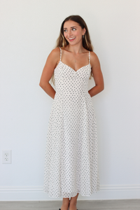 A woman modeling a white spaghetti-strap midi dress with a small black polka dot pattern, standing against a plain white wall.