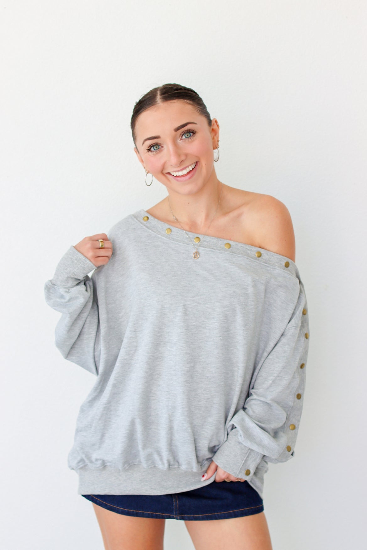 Woman wearing a light gray off the shoulder sweater with buttons along the trim 