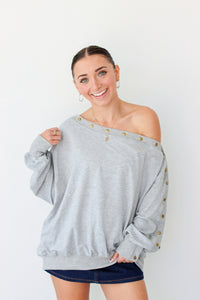 Woman wearing a light gray off the shoulder sweater with buttons along the trim 