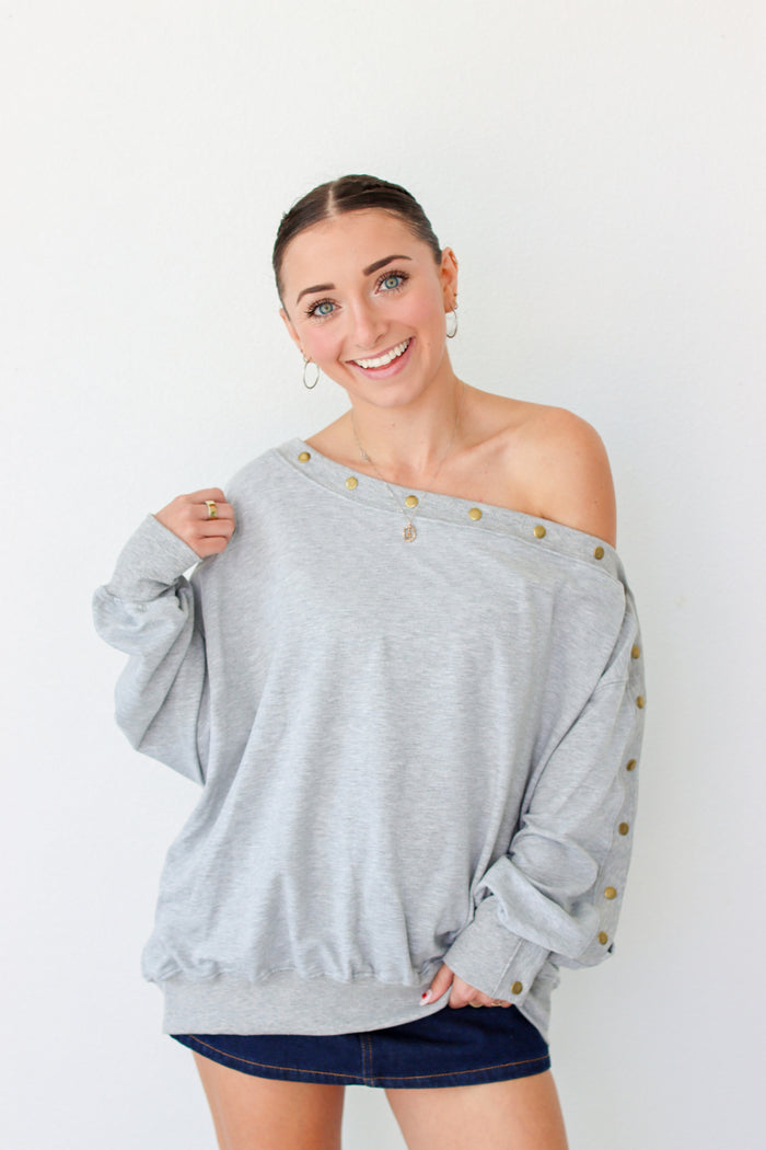Woman wearing a light gray off the shoulder sweater with buttons along the trim 