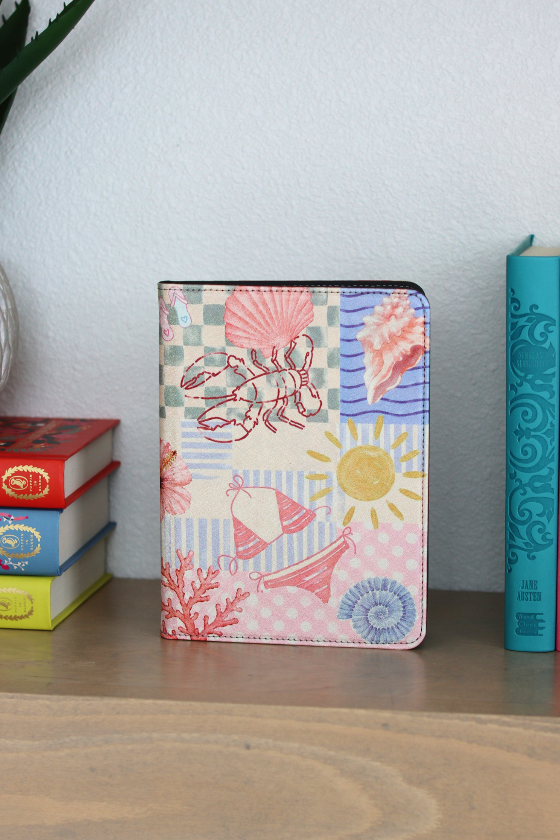 A kindle case with a colorful nautical patchwork print featuring a lobster, seashells, sun, and coral motifs, standing on a wooden surface next to books against a light wall.