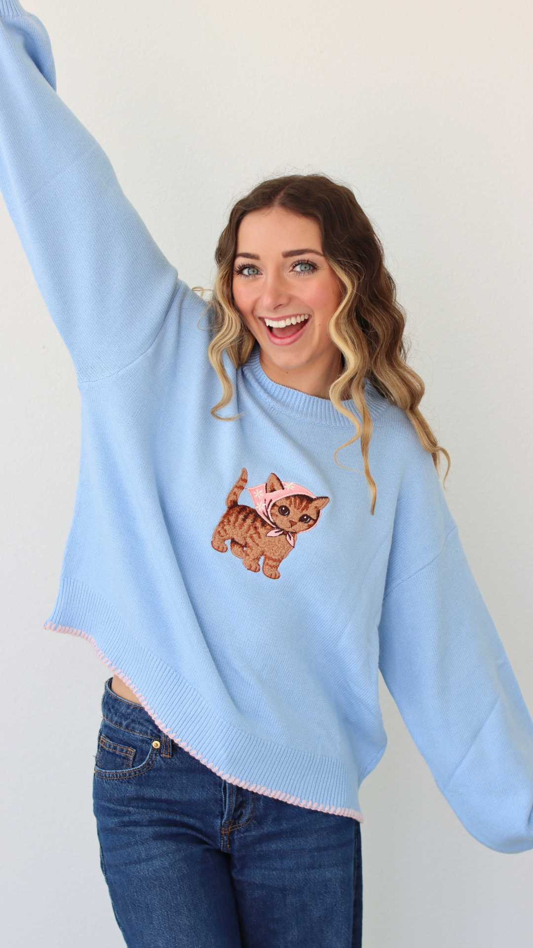 Woman wearing a light blue sweater with a cat embroidery design against a white background 
