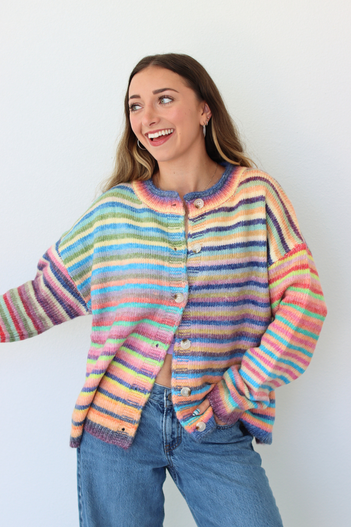 A woman wearing a colorful, fuzzy striped cardigan with wooden buttons and blue jeans, standing against a plain white wall.