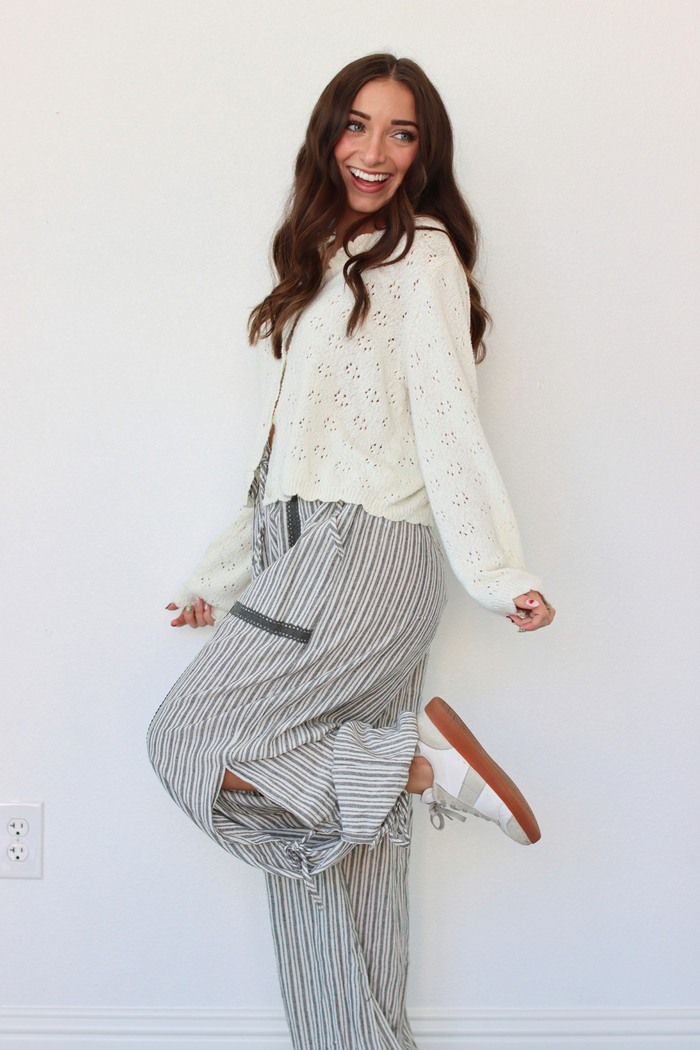 A woman modeling a cream eyelet knit cardigan over a white top and gray-and-white vertically striped wide-leg pants, standing against a plain white wall.