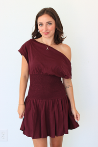 A woman modeling an off-the-shoulder mini dress in burgundy. The dress features an asymmetrical draped neckline, a smocked waist, and a flared skirt, shown against a plain white wall.