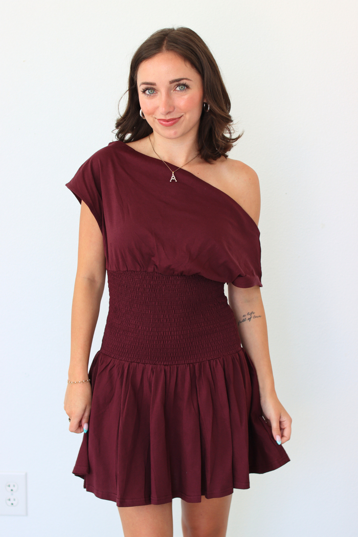 A woman modeling an off-the-shoulder mini dress in burgundy. The dress features an asymmetrical draped neckline, a smocked waist, and a flared skirt, shown against a plain white wall.