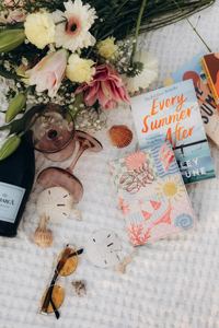 A styled flat-lay arrangement on a white textured blanket featuring a bouquet of pink, yellow, and white flowers, a dark wine bottle, two stemmed glasses, seashells, sunglasses, and Kindle Case