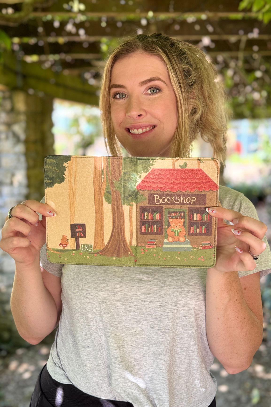 A woman holding a kindle case photographed outdoors under a pergola.