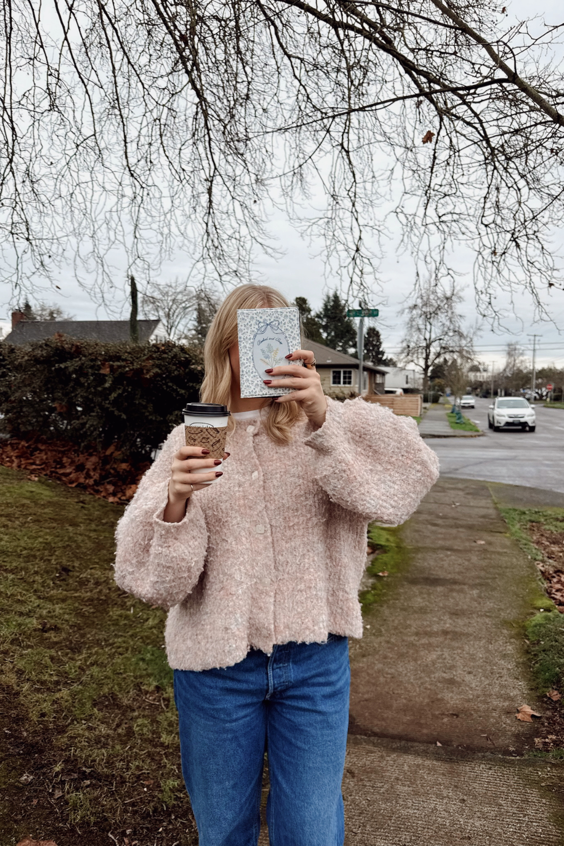 Person taking a mirror selfie outdoors, holding a coffee cup and a book.