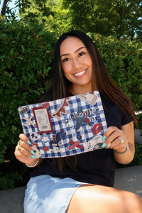 Woman holding a decorative book titled 'Chapter One' outdoors with greenery in the background