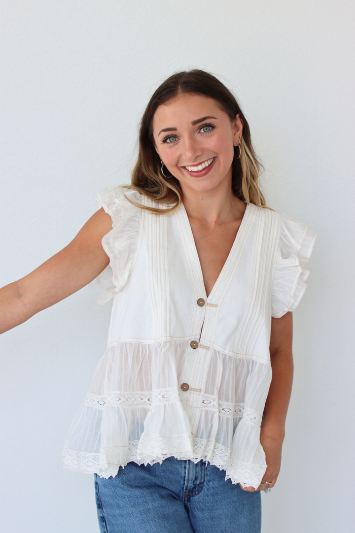 A woman wearing a white tiered blouse with ruffle sleeves and brown buttons, paired with blue jeans, standing against a plain white wall.