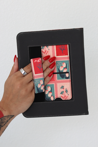A black faux-leather tablet sleeve with a pink and teal floral patterned strap inside, held by a hand with red nail polish against a plain white background.