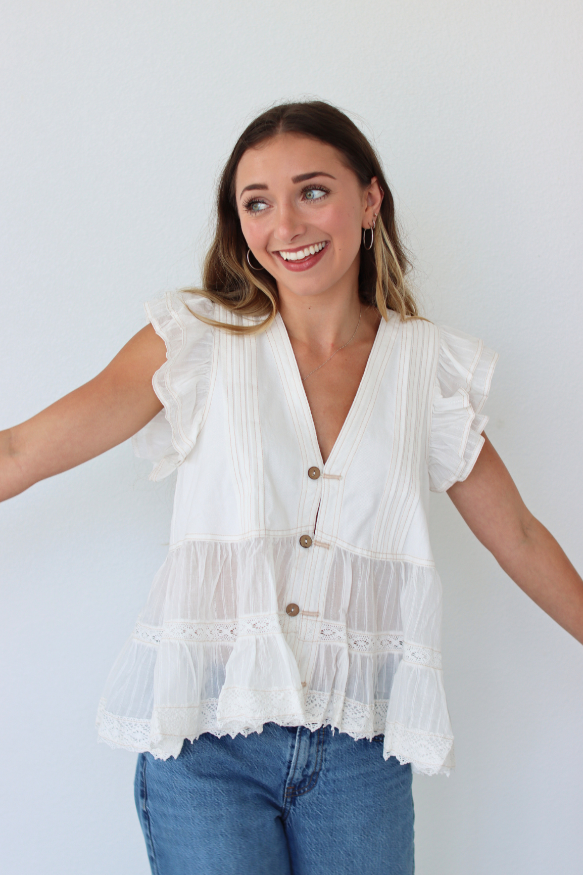 A woman wearing a white tiered blouse with ruffle sleeves and button details, paired with blue jeans, standing against a plain white wall.
