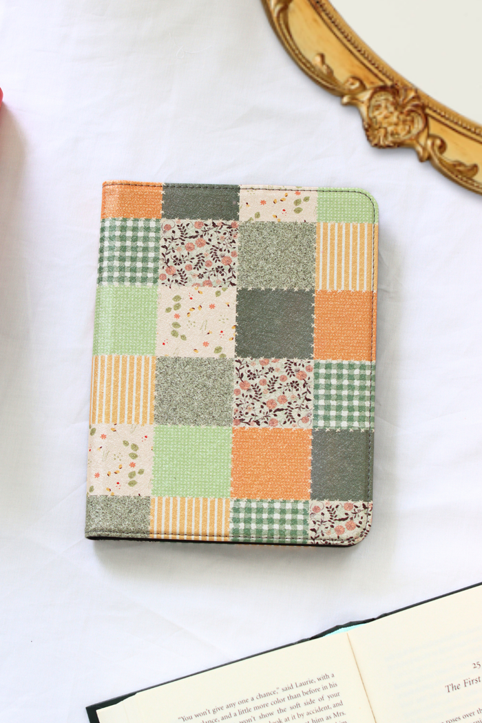 A square patchwork nook case with a multicolor design of green gingham, orange stripes, floral prints, and textured squares, resting on a white surface next to an open book and a gold mirror frame.