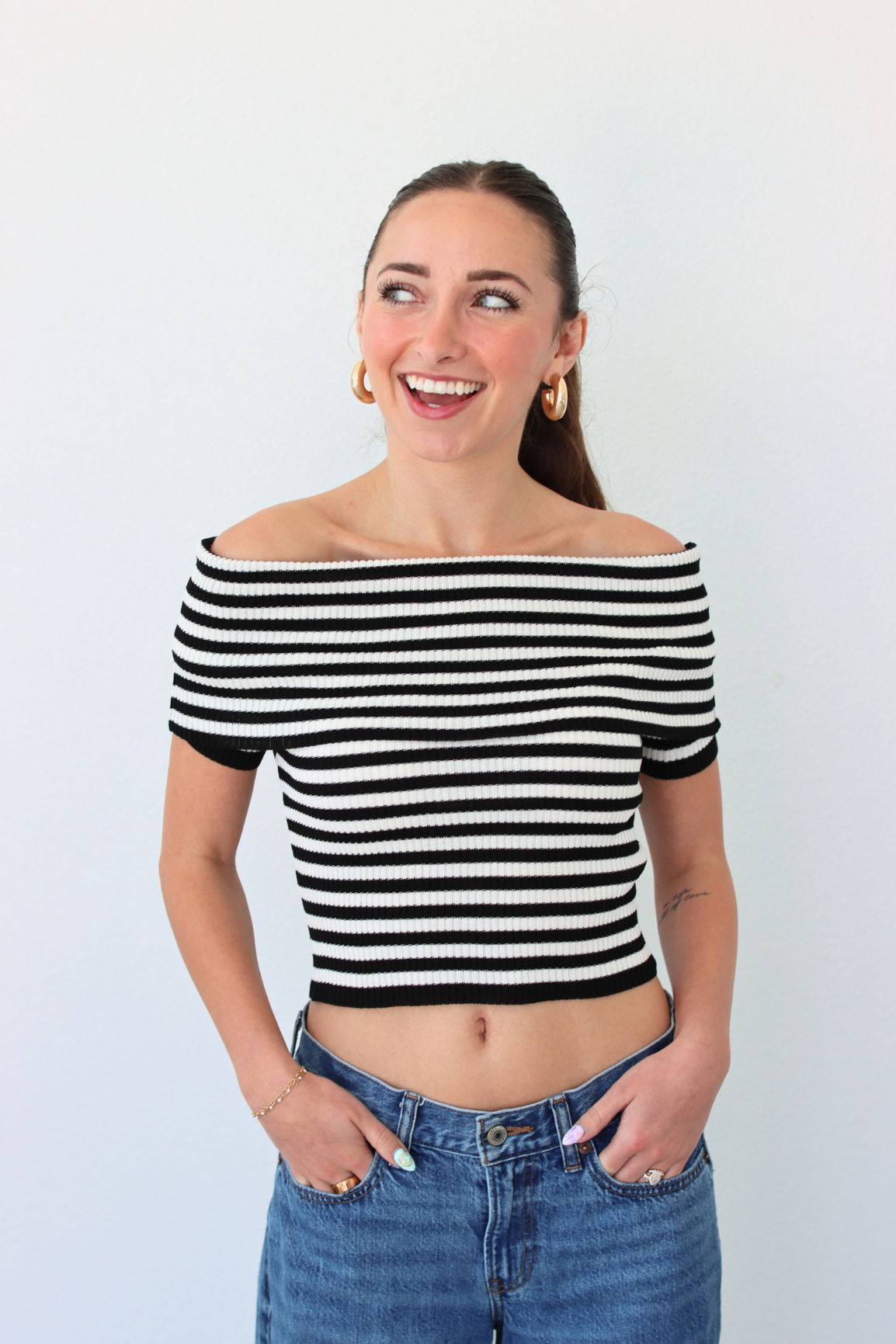 Woman wearing a black and white striped off-shoulder top and blue jeans against a plain background
